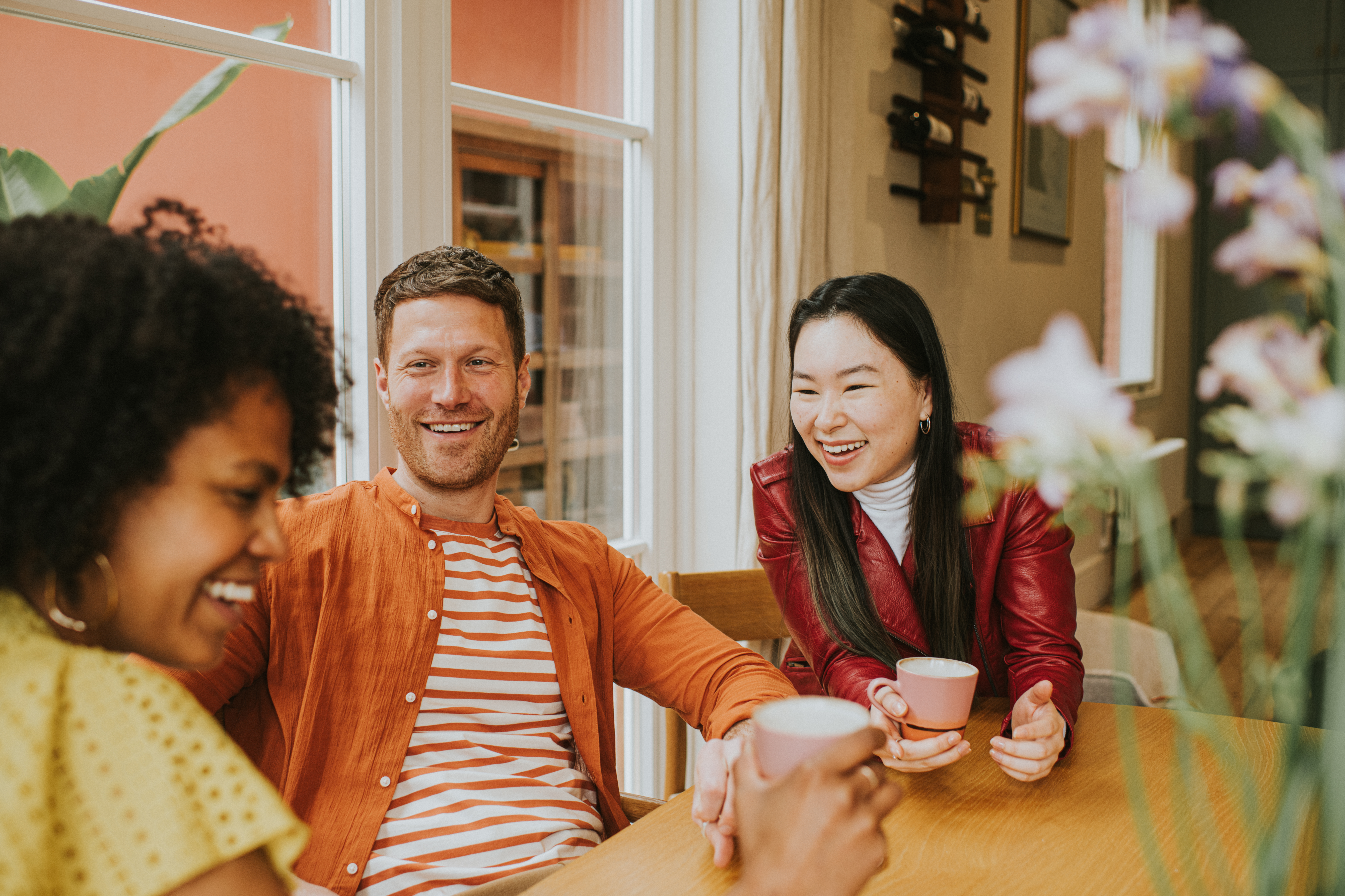 Three young people socialise at a table while enjoying hot drinks. They are animated as they giggle together. One girl is in the foreground whilst focus remains on a smiling male and female.